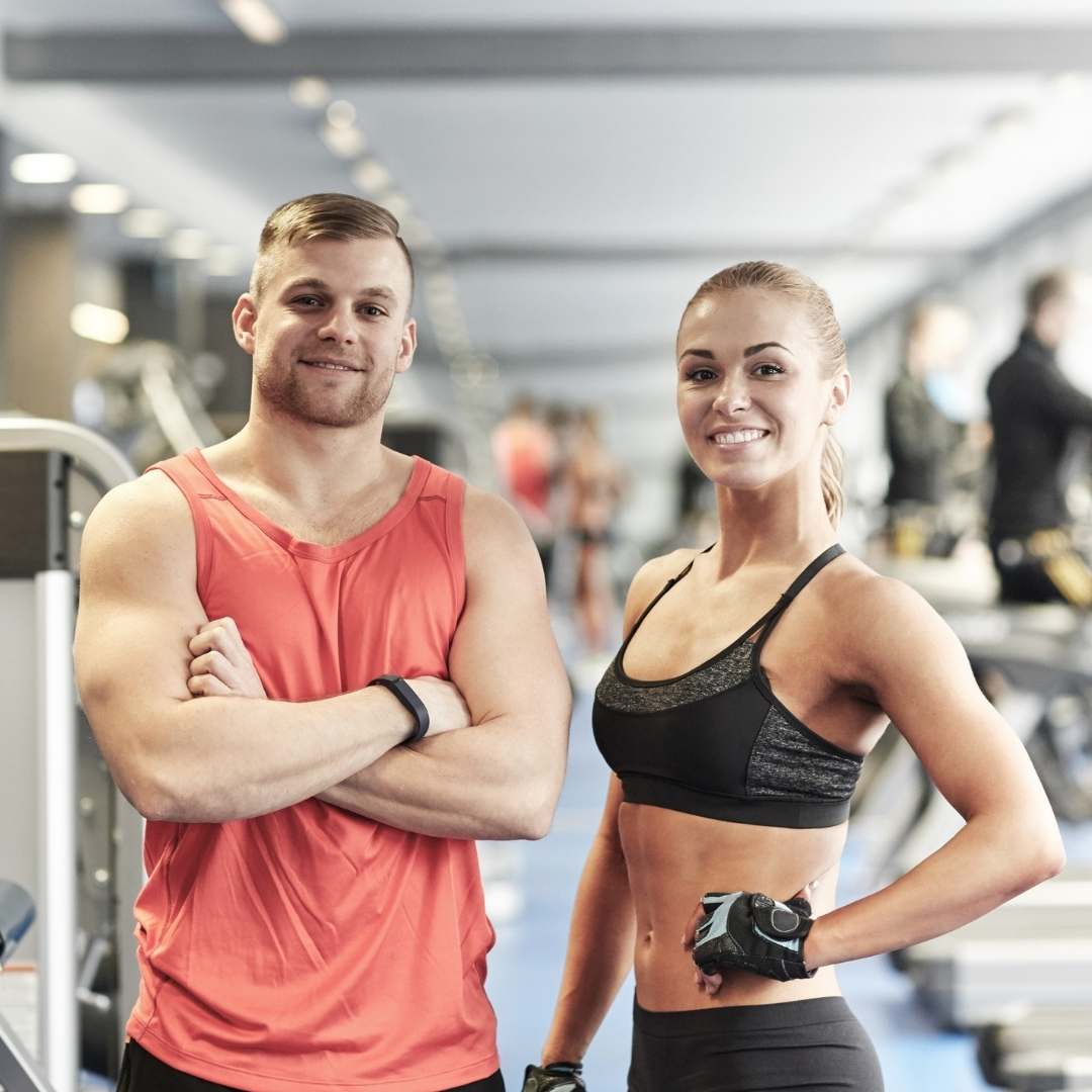 A man in a coral sleeveless athletic shirt and a woman in a black sports bra and leggings standing together in a brightly lit gym.