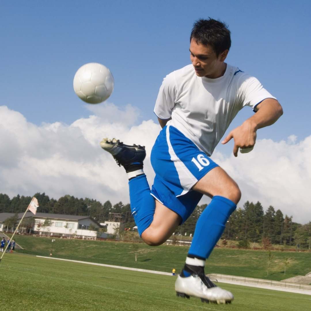 A soccer player in a white jersey and blue shorts with the number 16 kicks a white soccer ball on a green grass field under a blue sky with clouds.