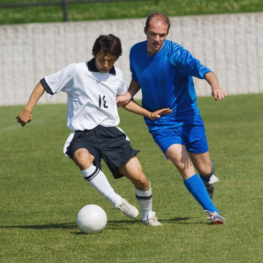 Two soccer players competing on a grass field, one wearing a white jersey and black shorts controlling a white soccer ball, and the other wearing a bright blue jersey and shorts.