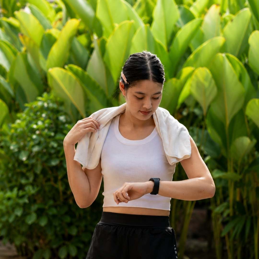A woman in athletic wear stands outdoors in front of lush green foliage, with a white towel draped over her shoulders. She is wearing a white sports bra or crop top, black athletic shorts, and a black smartwatch.