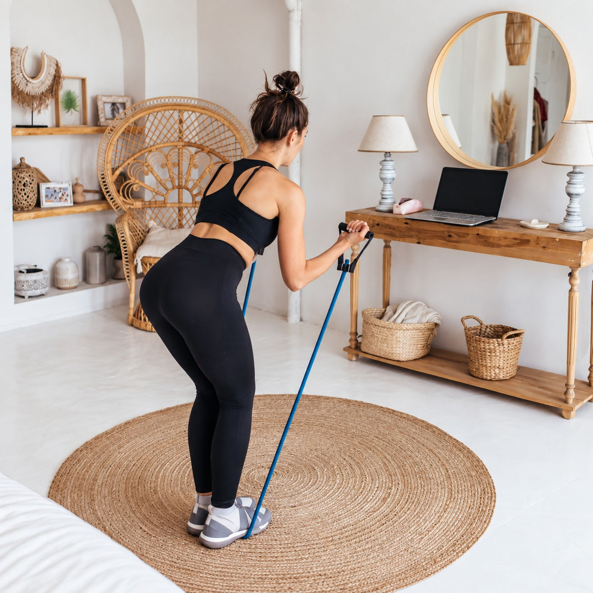 A woman in a black sports bra and leggings does resistance band exercises on a round jute rug in a bright, modern boho-style room.
