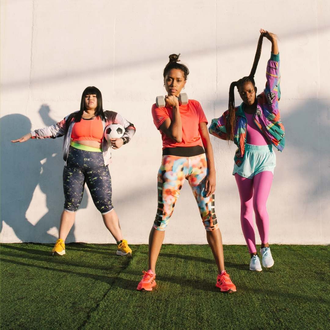 Three women posing in colorful athletic wear on artificial turf against a white wall, with strong sunlight casting shadows.